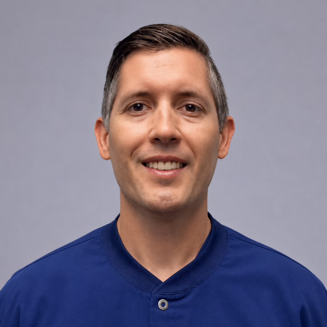 Smiling man with short dark hair in a blue shirt, portrait against a gray backdrop.