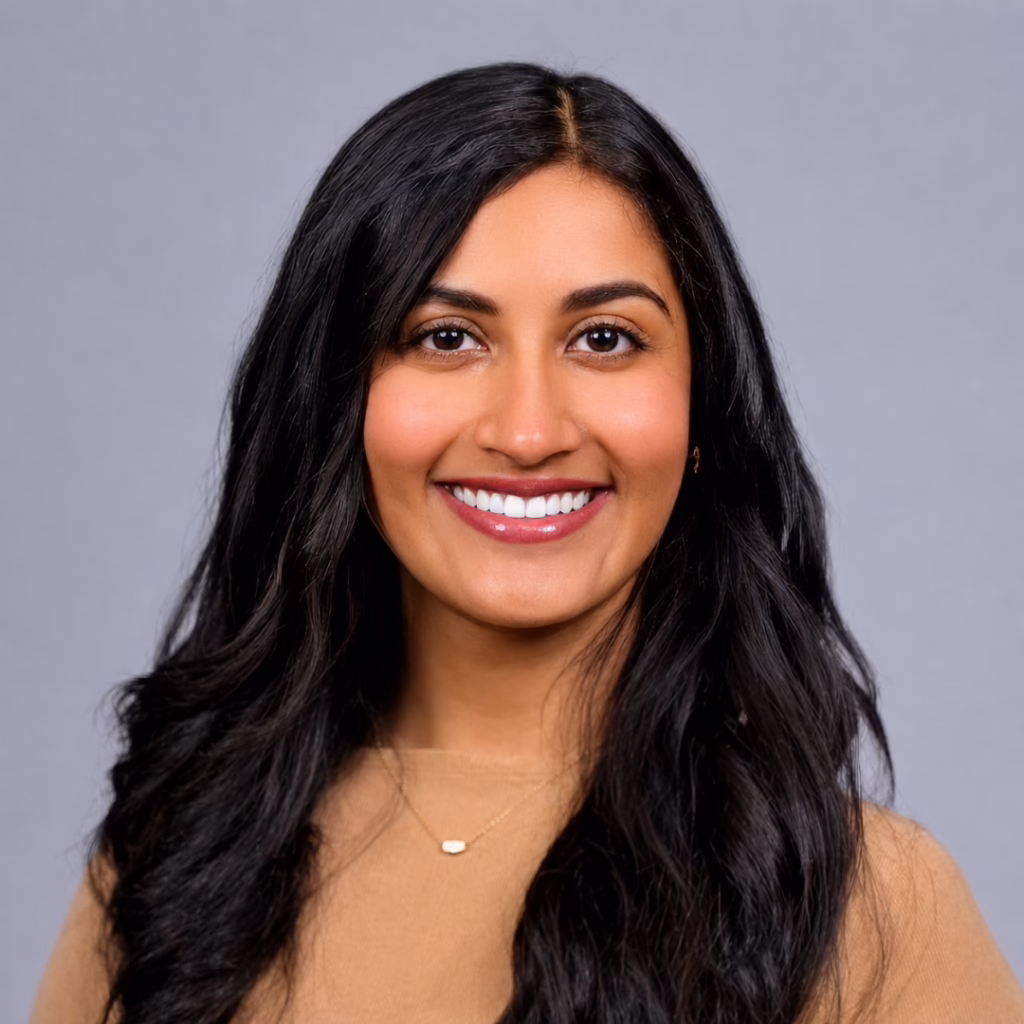 Head-and-shoulders portrait of a smiling woman with long black hair, wearing a beige top and a small gold necklace, against a gray background.